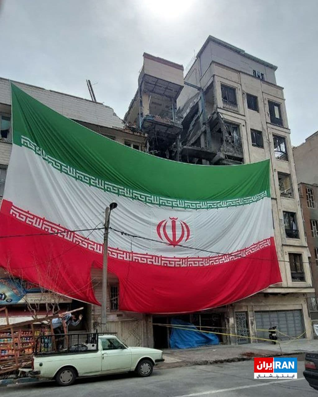 @ايران اينترنشنال: An image received by Iran International shows a demolished building in Tehran following airstrikes, which they have attempted to cover with an Islamic Republic flag.